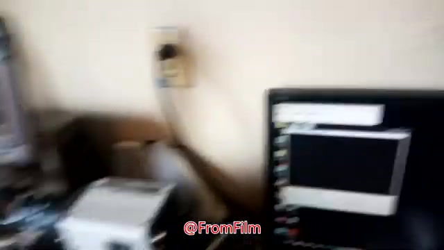 The still captures a workspace scene relevant to film-to-video transfer. A computer monitor displays software or tools used in the process, while a film projector sits nearby. The area appears cluttered, indicating a hands-on approach to the transferring process. Overall, it hints at the technical aspects involved in digitizing 16mm film.