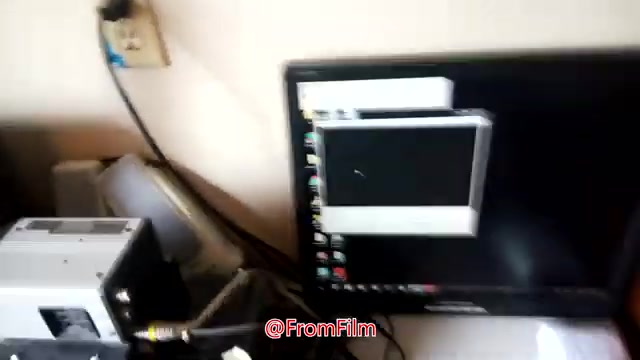 The still shows a cluttered workspace featuring a 16mm film projector positioned near a computer monitor. The monitor displays a video preview, indicating the film transfer process taking place. Cables connect the equipment, suggesting an active setup for digitizing film. The image captures a behind-the-scenes moment from a film-to-video transfer project by Brian Durham, highlighting the technical aspects of the transfer process.