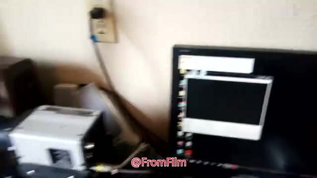 The still shows a cluttered workspace for film-to-video transfer. In the foreground, there's a film transfer device, alongside scattered papers and equipment. A computer monitor displays a blank screen, presumably part of the transfer setup. The environment appears informal, reflecting a hands-on approach to the process. The image captures an intimate glimpse into the behind-the-scenes work of film digitization.