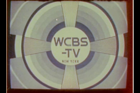 The still features the logo of WCBS-TV, presented within a circular design. The logo is centrally placed with 'WCBS' in bold letters above '-TV.' Surrounding it is a pattern of concentric circles and radiating lines in shades of blue and white. The text 'NEW YORK' is displayed below the logo. The overall aesthetic reflects the graphic design style of the early 1970s, characterized by bold colors and geometric shapes. The still is sourced from a 16mm negative derived from original 2-inch videotape.