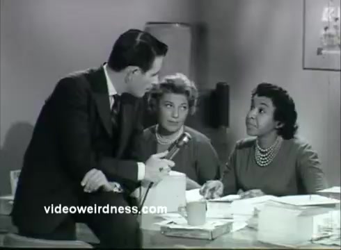 The still features a man in a suit interviewing two women at a desk. The setting appears to be an office, and the women are engaged in the conversation, with one looking directly at the interviewer. Papers and a microphone are present on the desk, suggesting a focus on information or discussion related to kidney disease awareness. The overall tone is formal and informative, characteristic of a public service announcement.