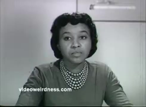 The still from the 'Kidney Disease Foundation of NY PSA' features a woman seated at a table, looking directly at the viewer. She has a serious expression and is wearing a simple, long-sleeved dress with a pearl necklace. The background suggests a domestic or professional setting, and the monochrome format adds a vintage quality to the image. This visual likely aims to convey a message about kidney health and awareness.