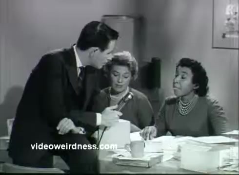 The still from the 'Kidney Disease Foundation of NY PSA' features a man in a suit interacting with two women seated at a desk. The women appear engaged in conversation, possibly discussing matters related to kidney health or fundraising. The setting looks like an office, with papers and documents scattered on the desk, highlighting a professional environment focused on health awareness.