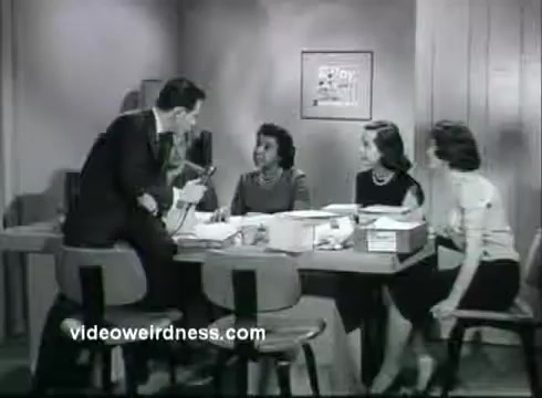 The still shows a 16mm black-and-white scene from a public service announcement (PSA) related to the Kidney Disease Foundation of NY. It features a man in a suit speaking to a woman at a table, while two other women sit nearby. The setting appears to be an office or meeting room, with paperwork and boxes on the table. The expression on their faces suggests a serious discussion, likely about kidney health and awareness.