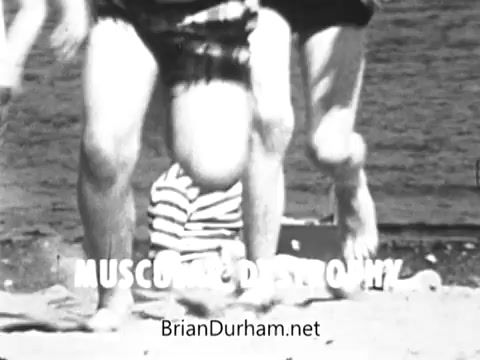 The still captures a moment from a Jerry Lewis Muscular Dystrophy PSA. It features a beach scene where two children, presumably playing or running, are shown in the foreground. The focus is on contrasting the vitality and movement of healthy children with the struggles faced by Spunky Hughes, a child with muscular dystrophy, who cannot join in these activities. The text 'MUSCULAR DYSTROPHY' is prominently displayed, underscoring the message of awareness and empathy.