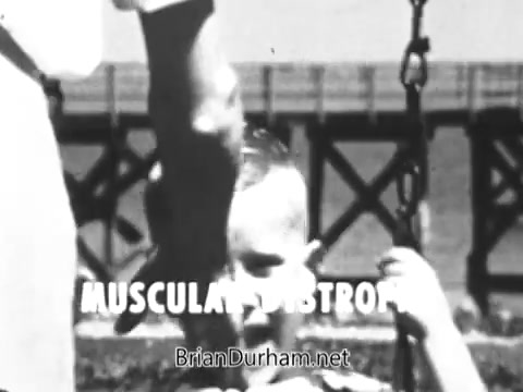 The still captures a poignant moment from the 'Jerry Lewis Muscular Dystrophy PSA' featuring a young boy, Spunky Hughes, on a swing. He appears to be struggling, emphasizing his condition of muscular dystrophy, which limits his ability to play independently. Nearby, an adult hand—presumably his father's—offers support, illustrating the bond and care between them. The text 'MUSCULAR DYSTROPHY' is prominently displayed, highlighting the focus of the PSA on raising awareness about the condition. The background suggests an outdoor setting, reinforcing a theme of love and protection in the face of adversity.
