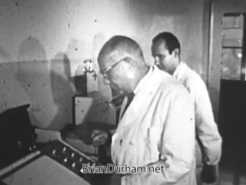 The still captures two men in lab coats, engaged in a scientific setting. One man is focused on a piece of equipment in front of him, while the other stands in the background, possibly observing or assisting. The environment appears clinical, with minimal decor, emphasizing the seriousness of their work. This scene likely connects to themes of research and effort related to muscular dystrophy, reflecting the emotional and physical struggles depicted in the PSA about Arnold Hughes and his son, Spunky.