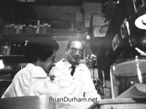 In the still from the 'Jerry Lewis Muscular Dystrophy PSA', two individuals, likely researchers, are engaged in discussion within a laboratory setting. One is in a white lab coat, with a thoughtful expression, while the other appears focused on their work. Shelves filled with books and scientific equipment surround them. The scene conveys a sense of dedication to the study of muscular dystrophy, reflecting the underlying theme of familial love and the pursuit of understanding and treatment for the condition.