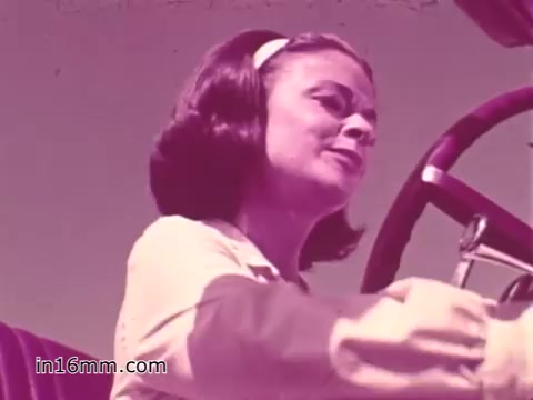 The still from the 1960s PSA features a young woman at the wheel of a vehicle, emphasizing themes of vehicle safety. She appears focused and responsible, reflecting the era's push for safe driving practices. The color palette has a vintage quality, typical of 16mm prints from that time. Overall, it conveys a message about the importance of safety in vehicle operation.