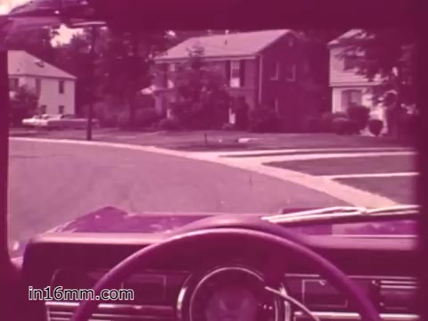The still captures a view from the driver's seat of a classic vehicle, focusing on the steering wheel and dashboard. In the background, suburban houses line a quiet street, indicative of 1960s America. The overall color tone has a vintage pinkish hue, typical of older film prints. This scene likely serves as a visual context for a public service announcement promoting vehicle safety in West Virginia during the 1960s.
