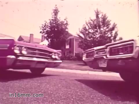 The still from the 16mm PSA showcases two classic vehicles parked side by side on a residential street, with a suburban house in the background. The color film has a slightly faded, pinkish hue typical of that era, reflecting the 1960s aesthetic. The emphasis appears to be on vehicle safety, likely highlighting the importance of car maintenance and inspections within the context of the public service announcement.