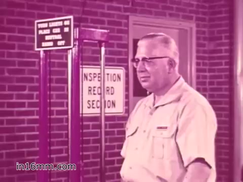 The still from the 16mm print of the 'West Virginia Safety Council PSA' features a man standing in front of a brick wall, near a vehicle inspection station. He appears to be discussing vehicle safety, likely emphasizing the importance of inspections. The signage nearby includes 'INSPECTION RECORD SECTION,' suggesting a focus on maintaining vehicle safety records. The overall tone is serious and informative, typical of public service announcements from the 1960s. The image likely has a vintage quality, indicative of its era.