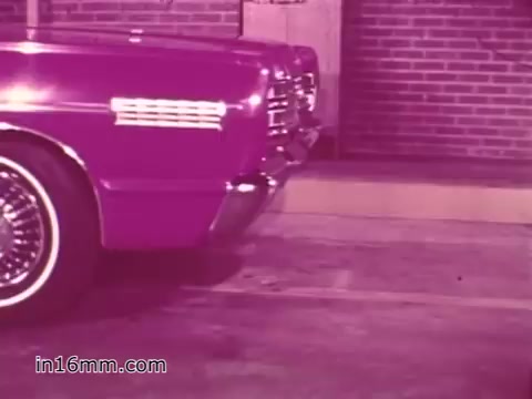The still from the 'West Virginia Safety Council PSA' features a vintage vehicle, likely from the 1960s, parked in front of a brick wall. The shot emphasizes the car's rear design, showcasing chrome details and wire wheels. The vibrant color palette, with a dominant purple hue, reflects the era's aesthetic. This scene is part of a public service announcement focused on vehicle safety and inspections.
