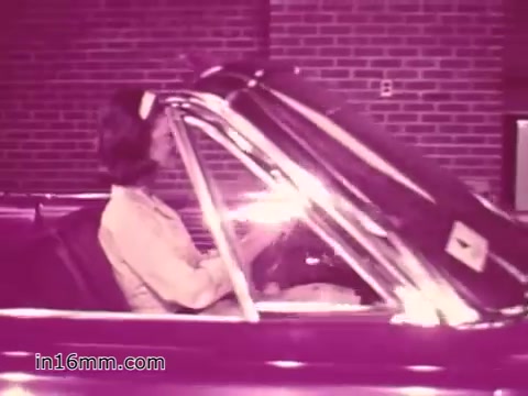 The still features a woman sitting in a convertible vehicle, likely during a safety inspection, in a setting with a brick wall backdrop. The image has a pinkish hue, typical of prints from the 1960s, emphasizing the focus on vehicle safety as part of a public service announcement by the West Virginia Safety Council.