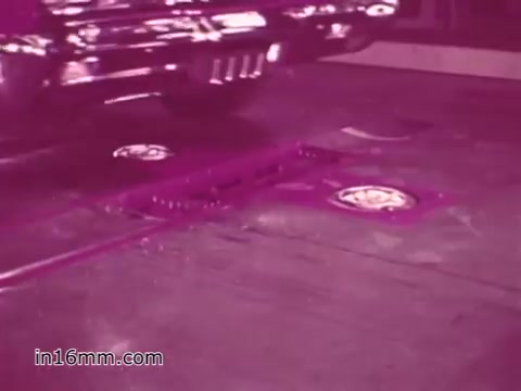 The still captures a vintage vehicle positioned over a safety inspection device, likely used to assess the car's condition. The scene is presented in a pinkish tint, typical of 1960s film prints. The focus appears to be on the vehicle's wheels, emphasizing safety inspection practices. The overall ambiance reflects the era's approach to road safety and public awareness.