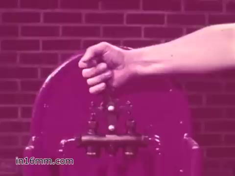 The still from the 16mm print of the 'West Virginia Safety Council PSA - 1960's' shows a close-up of a hand gripping a lever on a vehicle safety inspection tool. The background features brickwork, and the overall coloration has a vintage tone, typical of that era. This scene likely emphasizes the importance of mechanical safety checks in vehicles, aligning with the PSA's focus on promoting safe driving practices.