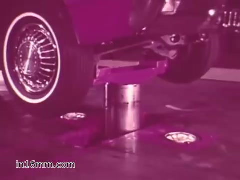 The still from the 'West Virginia Safety Council PSA' depicts a vintage vehicle raised above the ground on a hydraulic lift. The focus is on a wheel, showcasing its chrome detailing and tire. The background is infused with a pink hue, typical of the era's color grading in public service announcements. This image likely emphasizes vehicle safety and maintenance, reflecting the campaign's goal to promote safe driving practices.