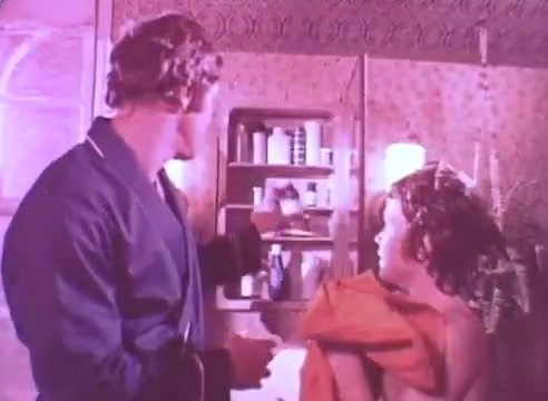 The still presents a scene from the film where a man in a robe is reaching for a bottle in a medicine cabinet, while a young girl stands beside him, wrapped in a towel. The setting appears intimate and domestic, emphasizing the importance of health and caregiving. The overall tone suggests that the moment is a reminder about taking blood pressure medication, framed within a familial context.