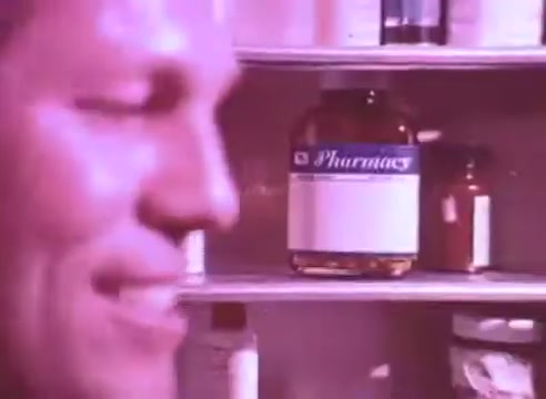 The still from 'Do it for them - take your blood pressure medication 4' features a smiling individual, possibly engaged in a positive or motivational moment. In the background, various medication bottles can be seen, including a prominently displayed container labeled 'Pharmacy.' The overall tone of the image suggests a focus on health and the importance of taking medication, emphasizing a supportive message. The color palette may lean towards a warm or pastel hue, enhancing the emotional appeal.