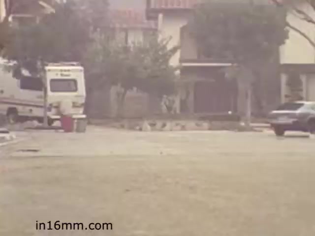 The still from 'Walk Safely,' an educational film from 1982, showcases a suburban residential scene. In the foreground, there is a grassy area with a parked RV and a car. The background features a neighborhood with houses and trees, portraying an everyday environment. The image has a soft, muted color tone typical of films from that time period, suggesting a focus on community awareness and safety in residential areas.