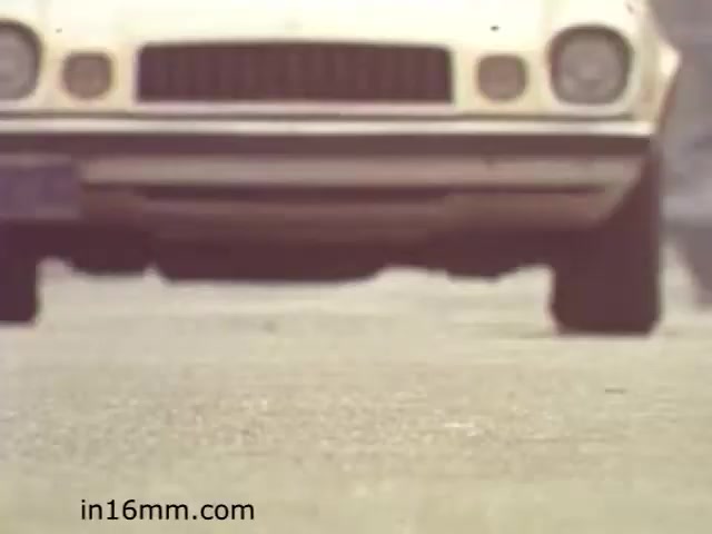 The still from the educational film 'Walk Safely' features a low-angle view of a white car approaching on a street. The focus is on the vehicle's front, highlighting its grille and bumper while the road surface is visible in the foreground. This framing creates a sense of perspective, possibly emphasizing the importance of being aware of traffic while walking. The film likely aims to educate viewers about pedestrian safety and awareness around vehicles.