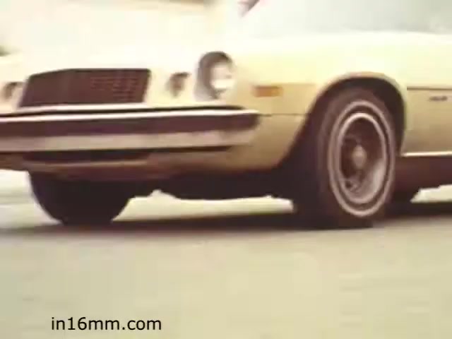 The still from 'Walk Safely,' an educational film from 1982 by Fiesta Films, features a close-up view of a vintage yellow car, likely highlighting themes of road safety. The image showcases the vehicle's front, including the grille and wheel, set against a blurred background that implies motion, suggesting the film may address topics related to pedestrian safety and traffic awareness. The aesthetic reflects the era's style, with the car's design typical of late '70s to early '80s vehicles.