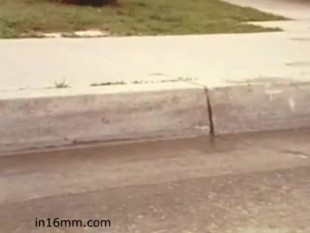 The 16mm still from 'Walk Safely,' an educational film from 1982 by Fiesta Films, depicts a close-up view of a sidewalk and curb. The concrete surface shows signs of wear, with a noticeable crack in the curb. The scene seems to emphasize pedestrian safety, possibly illustrating potential hazards for walkers. The context suggestive of teaching viewers to be aware of their surroundings while walking. The colors are typical of the era, possibly with a slightly muted palette.