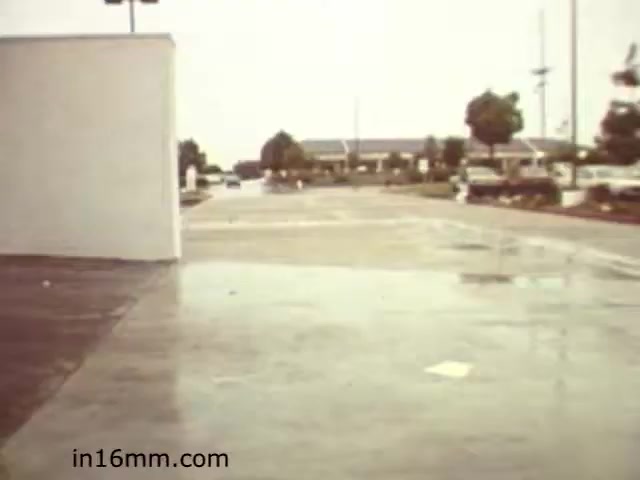 The still from 'Walk Safely,' an educational film from 1982 by Fiesta Films, depicts a rainy street scene. The foreground shows a wet pavement, while a white wall is visible on the left. In the background, you can see a commercial area with several cars and a few trees, indicating an urban environment. The overall atmosphere suggests a focus on pedestrian safety in suburban settings.