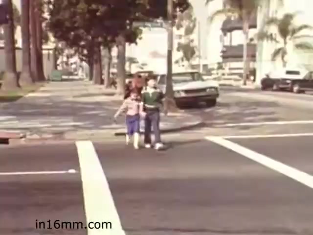 The still from 'Walk Safely,' an educational film from 1982, depicts two children standing at a crosswalk. They appear to be preparing to cross the street, highlighting themes of pedestrian safety. The setting features a tree-lined street with parked cars and urban infrastructure typical of the time. The scene likely emphasizes the importance of safe walking practices for children.