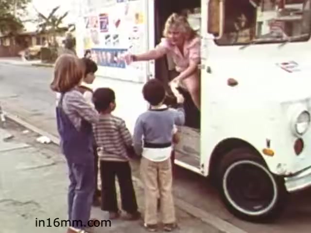 The still from 'Walk Safely,' an educational film from 1982 by Fiesta Films, depicts a scene with four children interacting with a woman in a white food truck. The children appear eager, likely anticipating a treat or snack. The setting looks like a suburban street, with houses and greenery in the background. The visual conveys a sense of curiosity and community, typical of educational films aimed at teaching safety and social interaction to children during that era.