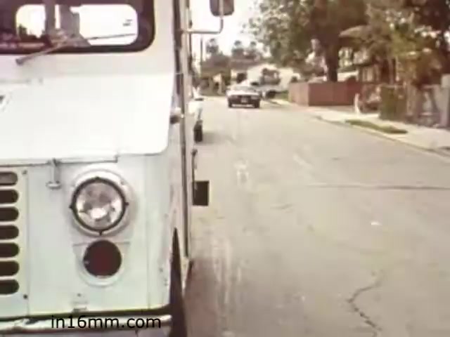 The still from 'Walk Safely,' an educational film from 1982, features a white truck on the left side of the frame, parked along a suburban street. The road appears quiet, with a few houses visible in the background. A car is seen driving in the distance, adding a sense of movement to the scene. The film likely addresses pedestrian safety, reflecting the era's focus on road awareness and safe walking practices. The visual aesthetic is characteristic of 1980s educational media, with a straightforward composition aimed at conveying its message clearly.
