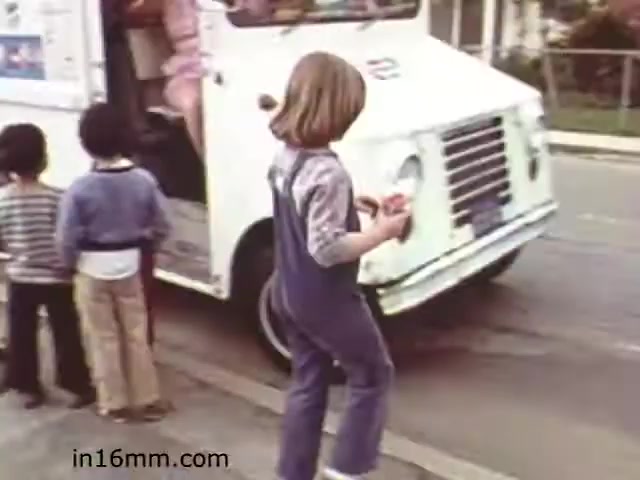 The still from 'Walk Safely,' an educational film from 1982, depicts a scene with children interacting near an ice cream truck. One child, dressed in a blue overall, stands in the foreground, looking at something in their hands, while two other children wait nearby. The ice cream truck appears to be parked, and the environment suggests a suburban neighborhood. This moment likely emphasizes pedestrian safety and awareness around vehicles, a key theme in the film.