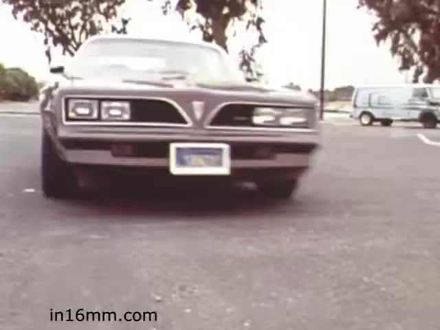 The still from 'Walk Safely - Educational Film from 1982 - Fiesta Films' features a close-up view of a gray Pontiac car parked in a lot. The vehicle is prominent in the foreground, and the shot emphasizes its front end, showcasing its hood and headlights. In the background, there are trees and another vehicle, indicating a suburban or semi-urban setting. The overall mood is reminiscent of the early 1980s, reflecting the style and aesthetics of that era. The film likely aims to promote safe walking practices, possibly highlighting the importance of awareness around vehicles.