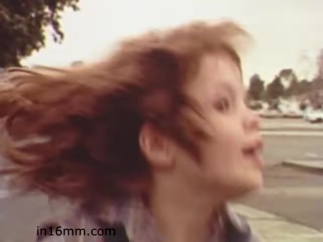 The still from 'Walk Safely,' an educational film from 1982 by Fiesta Films, shows a child with long hair in motion, suggesting an energetic and playful moment. The background appears to be a street scene, likely intended to emphasize safety while walking. The photograph captures a sense of spontaneity, typical of educational films aimed at teaching children about safe practices.