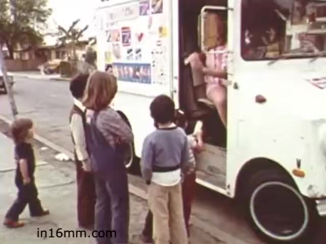 The still from 'Walk Safely,' an educational film from 1982, depicts a group of children gathered around an old-style ice cream truck. The truck is decorated with various stickers and advertisements, hinting at the nostalgic charm of that era. The kids, a mix of boys and girls, appear excited as they interact with someone inside the truck, likely waiting for treats. The scene conveys a sense of community and carefree childhood, typical of the time. This image captures a moment of joy and anticipation, likely reinforcing themes of safety and awareness in children's interactions with vehicles.