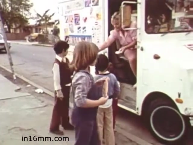 The still from 'Walk Safely,' an educational film from 1982 by Fiesta Films, depicts a group of children interacting with an ice cream truck. The scene captures a moment where a young blonde woman, likely the ice cream vendor, is handing an item to one of the children. The kids appear excited, showcasing a sense of innocence and joy typical of childhood experiences. The setting looks suburban, with a street view featuring trees and a sidewalk, highlighting communal interactions common in that era. Overall, it reflects themes of safety and community in a nostalgic, educational context.