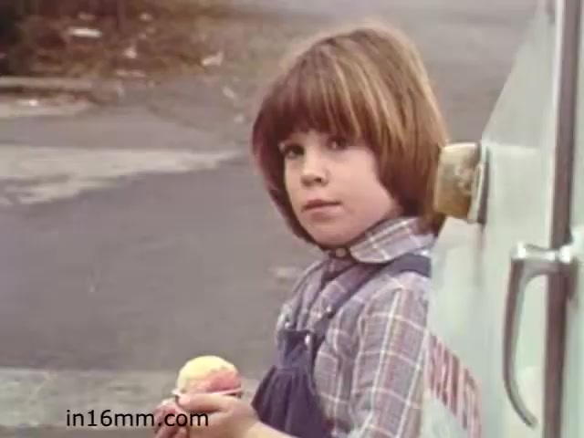 The still from the educational film 'Walk Safely' depicts a young child holding an ice cream cone while standing next to an ice cream truck. The child has a serious expression, with short brown hair and is wearing a plaid shirt and overalls. The background suggests a suburban setting, with a road and some greenery. This scene likely captures an everyday moment illustrating the theme of pedestrian safety, relevant to the film's educational purpose from the early 1980s.