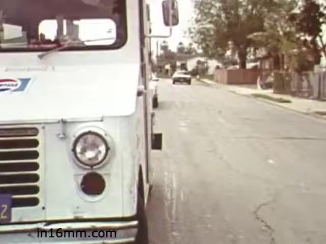The still from 'Walk Safely,' an educational film from 1982, features a vintage delivery truck positioned on an urban street. The truck is predominantly white with a faded logo, and its front is in focus, showcasing its distinctive grille and headlight. In the background, a car drives down the street amidst houses and greenery. The overall scene reflects a typical residential neighborhood from the early 1980s, capturing a moment likely intended to convey safety messages about pedestrian awareness and road safety.