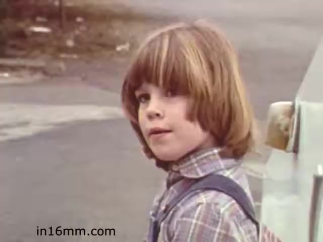 The still from the 1982 educational film 'Walk Safely' features a child with shoulder-length brown hair, wearing a plaid shirt and suspenders. The child appears to be looking back at something off-camera, with a thoughtful expression. The background suggests an outdoor setting, possibly a street or a park, typical of educational films aimed at teaching safety. The overall tone conveys a sense of innocence and awareness, fitting for the film's educational purpose.