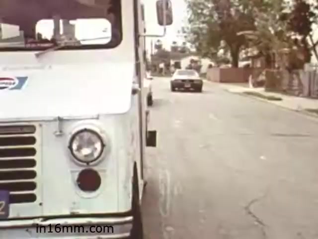 The still from 'Walk Safely,' an educational film from 1982 by Fiesta Films, features a street scene focused on a white delivery truck parked on the side of the road. In the background, a car is driving away, with residential buildings and trees visible. The overall setting appears to be suburban, indicative of the time period. The image likely emphasizes themes of road safety, relevant to pedestrian awareness or traffic education, typical of educational films from the era.