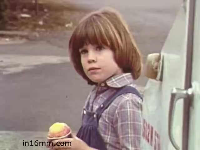 The still is from the educational film 'Walk Safely,' produced by Fiesta Films in 1982. It features a young girl with a bob haircut, dressed in a plaid shirt and blue overalls, holding an ice cream cone. She stands near an ice cream truck, with a slightly serious expression, suggesting a moment of reflection. The background hints at a suburban setting, emphasizing the film's focus on childhood safety awareness.