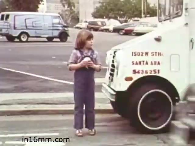The still from 'Walk Safely' depicts a young child standing on a sidewalk near a large delivery truck. The child appears to be holding something in their hands, possibly reflecting a moment of anticipation or caution. The truck is prominently featured, showcasing its wheel and part of the body, highlighting the importance of being aware of surroundings while near vehicles. This scene likely serves as a visual lesson on pedestrian safety, a common theme in educational films of that era. The background includes other vehicles and a cityscape, setting the typical urban environment for the film's message.