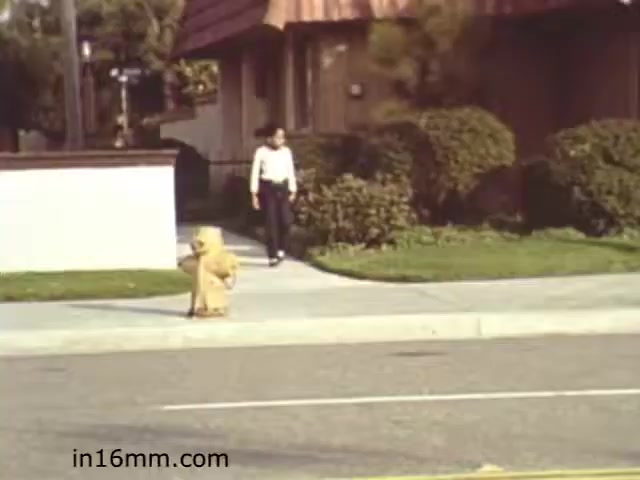 The still from the 1982 educational film 'Walk Safely' shows a sidewalk scene. A child, dressed in a yellow outfit, is seated on the curb, while an adult, wearing a white long-sleeve shirt, walks on the sidewalk nearby. The background features a residential area with greenery and a house, suggesting a suburban setting. The film likely aims to convey street safety messages, emphasizing awareness for both pedestrians and children.