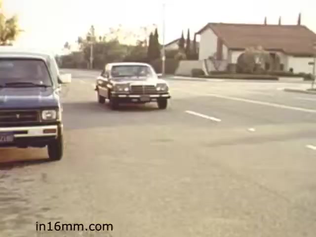 The still from 'Walk Safely' shows a street scene with a blue van parked on the left and a black car approaching from the right. The setting appears to be suburban, characterized by well-kept houses in the background and trees lining the street. The lighting suggests a late afternoon or early evening time. The film likely emphasizes pedestrian safety, focusing on traffic awareness and caution in urban environments.