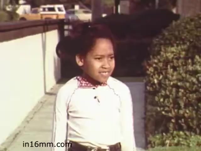 The still from 'Walk Safely,' an educational film from 1982, features a child standing on a sidewalk. The girl is dressed in a long-sleeved top with a contrasting collar and has her hair styled in a bun. The background shows a neatly maintained suburban street with a wall and some greenery, indicating a focus on pedestrian safety. The film likely aimed to educate children about safe walking practices in their environment.