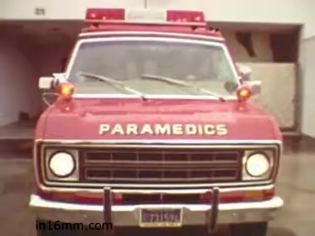 The still from the 1982 educational film 'Walk Safely' features a vintage red ambulance with the word 'PARAMEDICS' prominently displayed on its front. The vehicle is equipped with flashing lights on the roof, characteristic of emergency services. The background suggests a garage setting, and the image captures a moment that likely emphasizes the importance of safety and emergency response in the context of the film's educational message. This film appears to focus on promoting awareness around safety, possibly relating to pedestrian safety or emergency protocols.