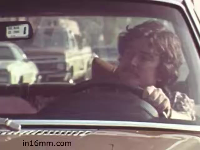 The still from 'Walk Safely,' an educational film from 1982 produced by Fiesta Films, depicts a man driving a car, featuring a vintage vehicle aesthetic characteristic of that era. His expression suggests a focus on the road, possibly conveying a message about safe driving practices. The background showcases other cars, hinting at an urban setting. The overall look is reminiscent of the 1980s, both in fashion and automotive design, reflecting the period's educational themes on safety.