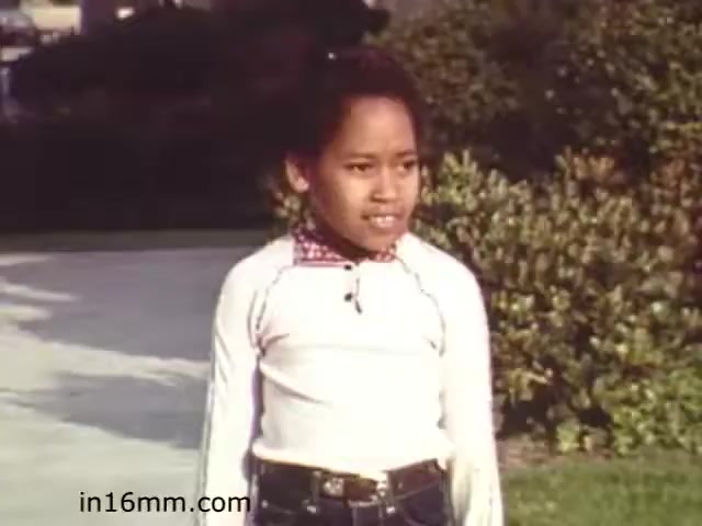 The still from 'Walk Safely,' an educational film from 1982, shows a young child standing outdoors, likely in a park or a similar setting. The child is wearing a white long-sleeve shirt and jeans, with a checkered collar visible. The background features greenery, suggesting a focus on safety in a natural environment. The film likely aims to educate viewers on pedestrian safety, aimed at children or families.