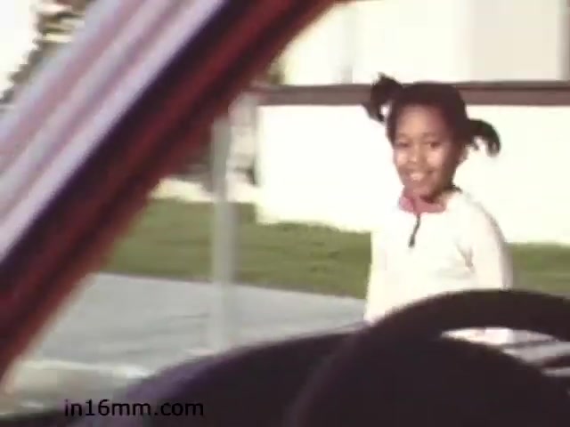 The still from the 1982 educational film 'Walk Safely - Educational Film from 1982 - Fiesta Films' depicts a young girl smiling while walking near a car. The image appears to be shot from inside a vehicle, giving a perspective of the view from the driver’s seat. The background includes green grass and a building, suggesting an outdoor setting that emphasizes themes related to pedestrian safety and awareness. Overall, the scene conveys an educational message likely aimed at promoting safety while walking.