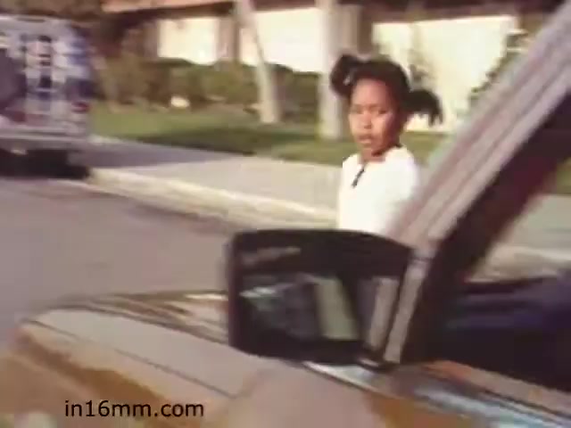 The still from 'Walk Safely,' an educational film from 1982, shows a young girl walking near a street, seemingly unaware of an approaching vehicle. The scene emphasizes pedestrian safety, typical of educational films aimed at teaching children to be cautious around traffic. The setting appears suburban, with houses and parked vehicles in the background. The girl's expression suggests a moment of focus or mild concern, aligning with the film's theme of promoting awareness and safety.